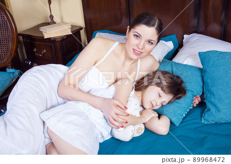 Mother looking at camera with toothy smiling and her daughter sleeping near. Studio shot Mother looking at camera with toothy smiling and her daughter sleeping near. Studio shot 89968472