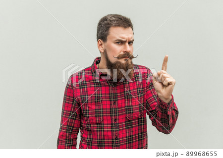 Warning sign. Young adult businessman with beard and handlebar mustache looking at camera with serious face and finger warning. Studio shot, on gray background. Warning sign. Young adult businessman with beard and handlebar mustache looking at camera with serious face and finger warning. Studio shot, on gray background. 89968655
