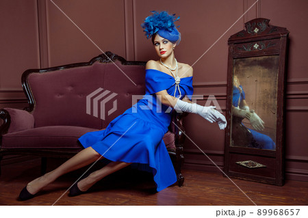 Old Fashioned retro great britain style photography. Beautiful young caucasian model in blue dress and fashion makeup and hat posing sitting on the artificial sofa and looking into the distance. 89968657