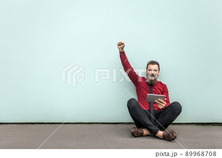Working in outdoor, sits on the ground. Young adult bearded businessman work in tablet, hand up and shout with smile. Outdoor shot on gray wall. Working in outdoor, sits on the ground. Young adult bearded businessman work in tablet, hand up and shout with smile. Outdoor shot on gray wall. 89968708