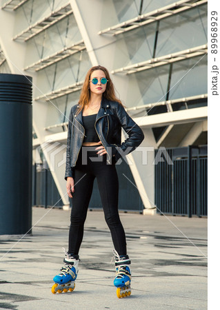 Girl on rollerblades standing in building background. Young fit women girl in blue sunglasses, jeans and jacket on roller skates riding outdoors after rain. 89968929