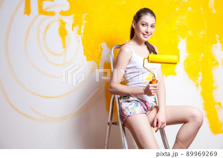 Beautiful young caucasian model in coloured shorts and white shirt and posing holding outrigger in her hands, painting the wall and looking at camera. Studio shot. 89969269