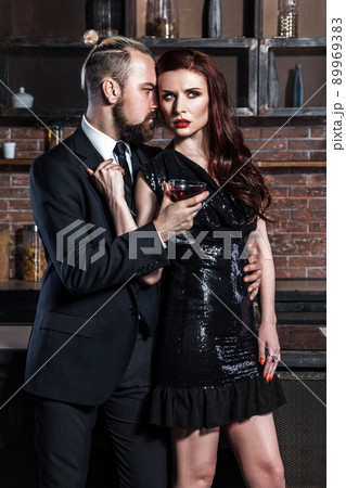 Well dressed famous couple in loft interior. Man in classic style and tie, woman in black dress. Couple holding glasses of vine. Studio shot, brick wall Well dressed famous couple in loft interior. Man in classic style and tie, woman in black dress. Couple holding glasses of vine. Studio shot, brick wall 89969383