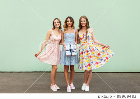 Charming three beautiful young adult friends with dresses and present box smiling and looking at camera. Isolated on green wall. Studio shot 89969482