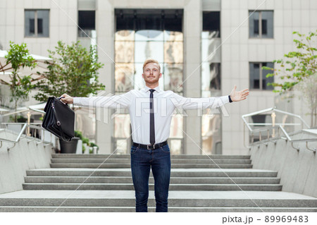 Celebrating success. Young adult red hair and bearded businessman keeping arms raised and expressing positivity while standing outdoors with office building in the background Celebrating success. Young adult red hair and bearded businessman keeping arms raised and expressing positivity while standing outdoors with office building in the background 89969483