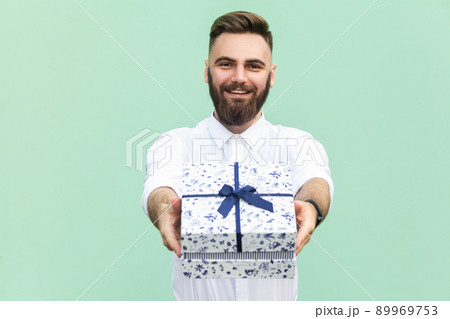 Businessman holding gift box and looking at camera and toothy smile. On light green background. Studio shot 89969753