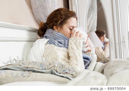 Sick woman with cup of tea. Closeup image of young frustrated woman in knitted blue scarf holding and drinking a cup of tea while sitting in bed of her room. 89970004
