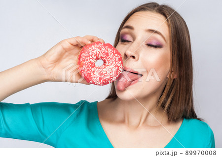 Portrait of pleasure dreaming young beautiful girl in blue blouse standing, holding and showing pink donut, trying to suck with tongue out in grey background. Indoor, isolated, copy space, studio shot Portrait of pleasure dreaming young beautiful girl in blue blouse standing, holding and showing pink donut, trying to suck with tongue out in grey background. Indoor, isolated, copy space, studio shot 89970008