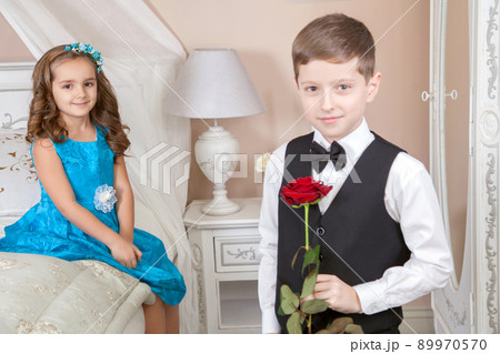 Beautiful young prince in tuxedo suit and princess holding rose in blue dress and fashion band posing sitting on the bed in bright room and looking into the distance. Love story. Studio shot. Beautiful young prince in tuxedo suit and princess holding rose in blue dress and fashion band posing sitting on the bed in bright room and looking into the distance. Love story. Studio shot. 89970570
