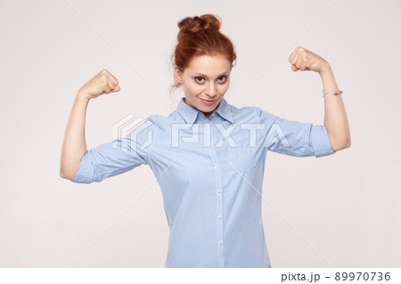 Beautifulwoman posin looks like bodybuilder, showing biceps, looking at camera and smiling. Indoor. Gray background 89970736