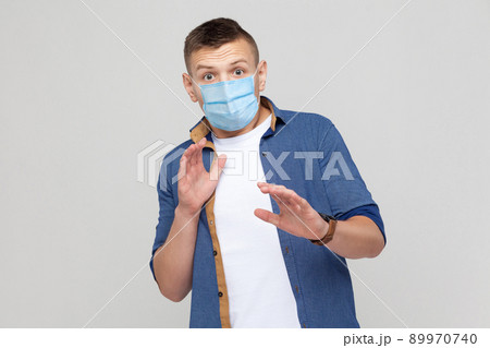 No, wait. Portrait of scared young man in casual style with surgical medical mask standing and looking at camera, afraid and panic. indoor studio shot, isolated on gray background. No, wait. Portrait of scared young man in casual style with surgical medical mask standing and looking at camera, afraid and panic. indoor studio shot, isolated on gray background. 89970740