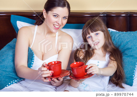 Happy mother and daughter having fun and holding cup and resting in bed, looking at camera with toothy smile. 89970744