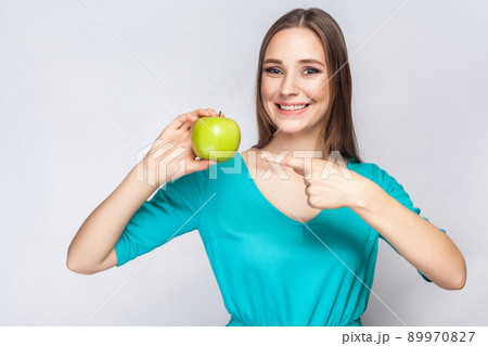 Young beautiful woman with freckles and green dress holding apple and pointing with finger. studio shot, isolated on light gray background. 89970827