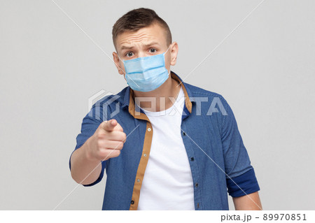 Hey you, are you serious?. Portrait of serious young man in casual style with surgical medical mask standing, frowning and poinitng at camera . indoor studio shot, isolated on gray background. Hey you, are you serious?. Portrait of serious young man in casual style with surgical medical mask standing, frowning and poinitng at camera . indoor studio shot, isolated on gray background. 89970851