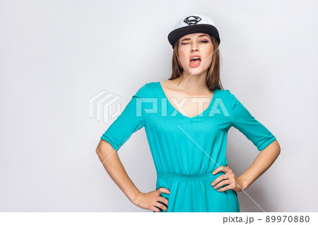 Portrait of beautiful young girl in blue dress and white cap with supermen sign standing, holding hands on waist, sneezing and winking with open mouth. Indoor, studio shot, copy space, grey background 89970880