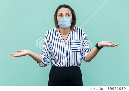 I don't know what to do. Portrait of confused middle aged woman with surgical medical mask standing with raised arms and thinking. medicine, health care concept. indoor, isolated on blue background. I don't know what to do. Portrait of confused middle aged woman with surgical medical mask standing with raised arms and thinking. medicine, health care concept. indoor, isolated on blue background. 89971080
