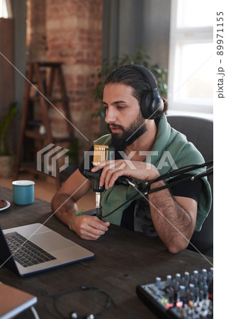 Vertical medium portrait shot of stylish Middle Eastern man with beard on face sitting at desk making content for video blog 89971155