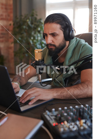 Vertical medium portrait of serious handsome man wearing headphones sitting at desk typing something on laptop 89971160
