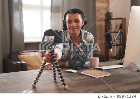 Medium portrait of beautiful young female blogger wearing headphones sitting at desk in loft room fixing camera on tripod 89971234