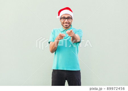 Hey you! Its christmas man! Bearded man in santa hat and glasses, pointing finger and looking at camera with toothy smile. On gray background. Indoor, studio shot Hey you! Its christmas man! Bearded man in santa hat and glasses, pointing finger and looking at camera with toothy smile. On gray background. Indoor, studio shot 89972456