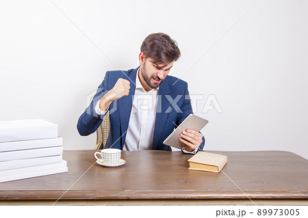 Technology, people and business concept - handsome man with beard and brown hair and blue suit and tablet pc computer and some books sitting in the office angry and unhappy. .Isolated on white Technology, people and business concept - handsome man with beard and brown hair and blue suit and tablet pc computer and some books sitting in the office angry and unhappy. .Isolated on white 89973005