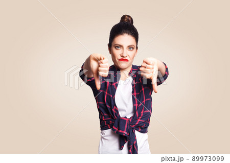 Dislike. Young unhappy upset girl with casual style and bun hair thumbs down her finger, on beige blank wall with copy space looking at camera with toothy smile. focus on face. 89973099