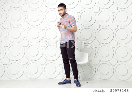 Young bearded fashion model in casual style is posing near white circle wall background. studio shot. looking at camera with pointing finger. 89973148