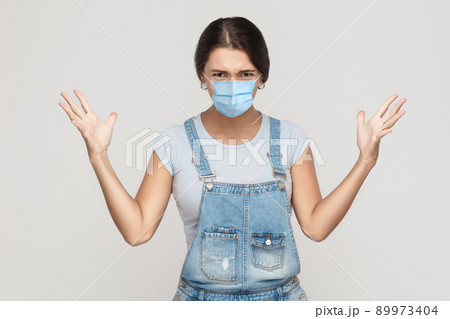 What? Portrait of shocked crazy young woman with surgical medical mask in denim overalls standing, raised arms and looking at camera with angry face. indoor studio shot isolated on gray background. 89973404