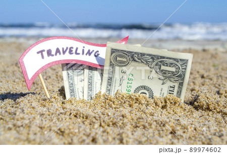 Paper bills one dollar and small stick with paper speech bubble with word Traveling buried in sand beach background sea close-up sunny summer day. Concept Money travel tourism vacation holiday relax. Paper bills one dollar and small stick with paper speech bubble with word Traveling buried in sand beach background sea close-up sunny summer day. Concept Money travel tourism vacation holiday relax. 89974602