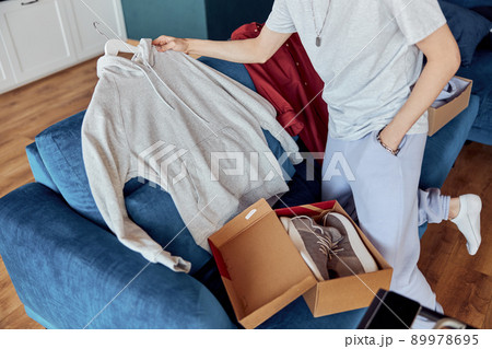 Young man unpacking online shopping at home while holding hoodie 89978695