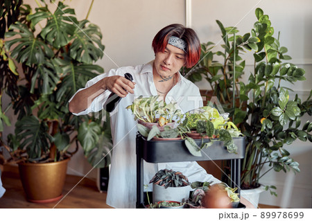 Asian guy spraying flowerpots on a stand in the room 89978699