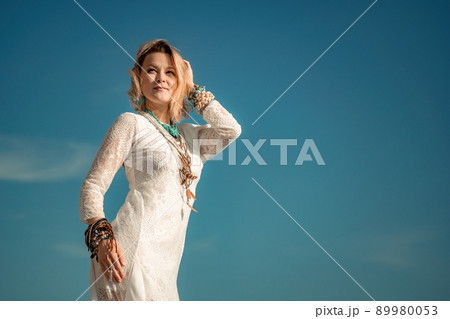 Woman in bohemian clothing on beach at sunset. Boho style for fashionable look on resort. Middle aged well looking woman in white dress and boho style braclets. Summer fashion Woman in bohemian clothing on beach at sunset. Boho style for fashionable look on resort. Middle aged well looking woman in white dress and boho style braclets. Summer fashion 89980053