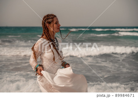 Model in boho style in a white long dress and silver jewelry on the beach. Her hair is braided, and there are many bracelets on her arms. Model in boho style in a white long dress and silver jewelry on the beach. Her hair is braided, and there are many bracelets on her arms. 89984671