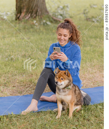 woman with a dog in the park does sports or fitness on a yoga mat. a European woman with curly hair in a blue hoodie is chatting on a smartphone 89987690