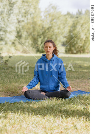 woman in the park does yoga, sports, meditation or fitness on a yoga mat. a European woman with curly hair in a blue hoodie takes care of her body and mental health woman in the park does yoga, sports, meditation or fitness on a yoga mat. a European woman with curly hair in a blue hoodie takes care of her body and mental health 89987691