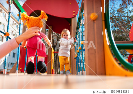 A cute little baby is playing on a children's slide. A woman's hand holds out a doll for a girl. The concept of child development and happy childhood A cute little baby is playing on a children's slide. A woman's hand holds out a doll for a girl. The concept of child development and happy childhood 89991885