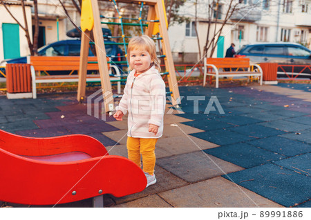 A little cute girl is standing near the children's slide on the playground. The concept of childhood and autism A little cute girl is standing near the children's slide on the playground. The concept of childhood and autism 89991886