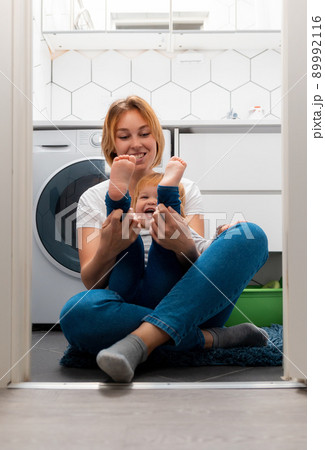 Happy mother and her little daughter play together in the bathroom, sitting on the floor. Vertical. Washing machine in the background 89992116