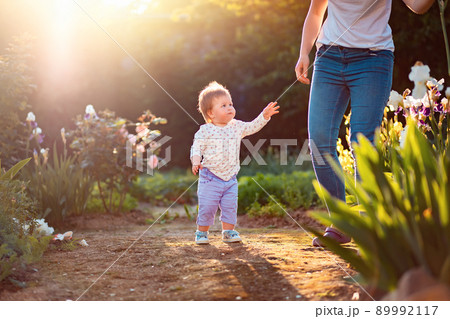 A cute little kid looks at his mom and reaches out to her. In the background, a summer garden and flowering shrubs. Happy children's day 89992117