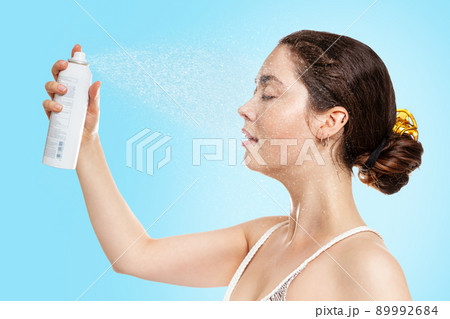 A young Caucasian woman with wet skin sprays thermal water on her face. Blue background. Side view. The concept of cosmetology, skin care and moisturizing A young Caucasian woman with wet skin sprays thermal water on her face. Blue background. Side view. The concept of cosmetology, skin care and moisturizing 89992684