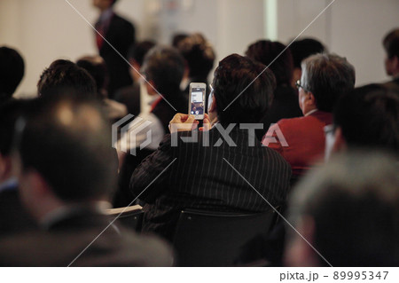 会議場でイスに座ってスマートフォンで写真を撮る男性の後ろ姿 89995347