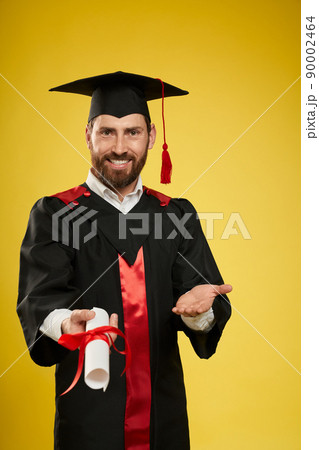 Front view of young male with beard graduating from college. Boy in graduate gown and mortarboard standing, showing diploma, smiling. Isolated on yellow studio background, Front view of young male with beard graduating from college. Boy in graduate gown and mortarboard standing, showing diploma, smiling. Isolated on yellow studio background, 90002464