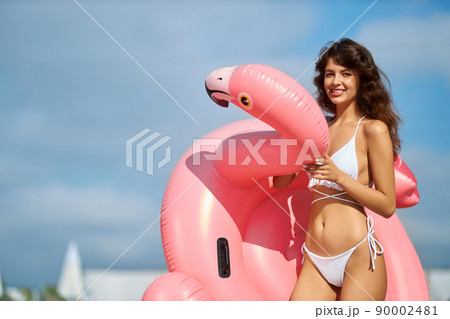 Charming slim lady walking with inflatable coral flamingo, looking at camera at sunny day. Front view of female model holding funny pink bird against of blurred skyline. Concept of water activity. 90002481