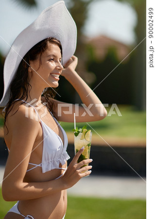 Smiling female in bikini and big hat standing with glass of cool mojito outdoor. Side view of cheerful long haired girl drinking cocktail with slices of lemon while enjoying vacation. Concept of rest. 90002499