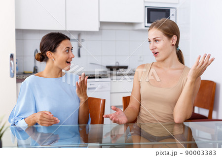 Young smiling emotional girlfriends sharing secrets and sitting at table 90003383