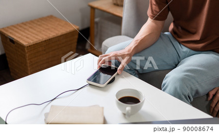 Technology Concept A person with his light blue jeans sitting on the couch and trying to charge his smartphone on the wireless battery charger 90008674