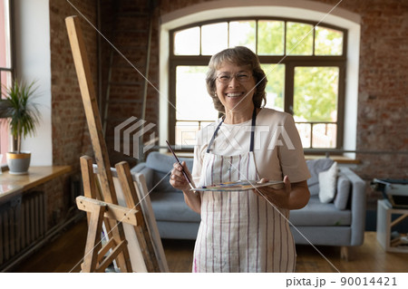 Older painter woman posing near easel with palette and paintbrush Older painter woman posing near easel with palette and paintbrush 90014421