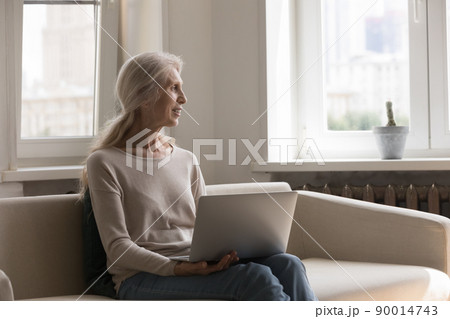 Happy pensive elder 70s woman holding laptop, resting on couch 90014743