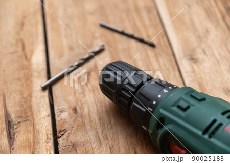 An electric drill and two drills lie against the wooden surface. The hand-held power tool is turning the boards. Selective focus. No people. An electric drill and two drills lie against the wooden surface. The hand-held power tool is turning the boards. Selective focus. No people. 90025183