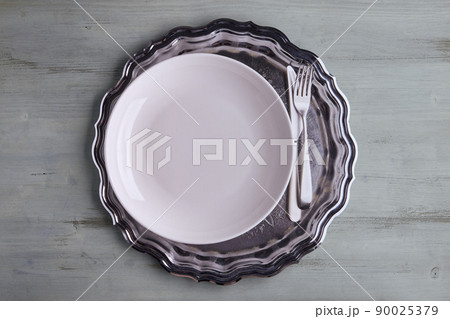 A white plate with cutlery on a shiny metal tray on a light wooden table 90025379